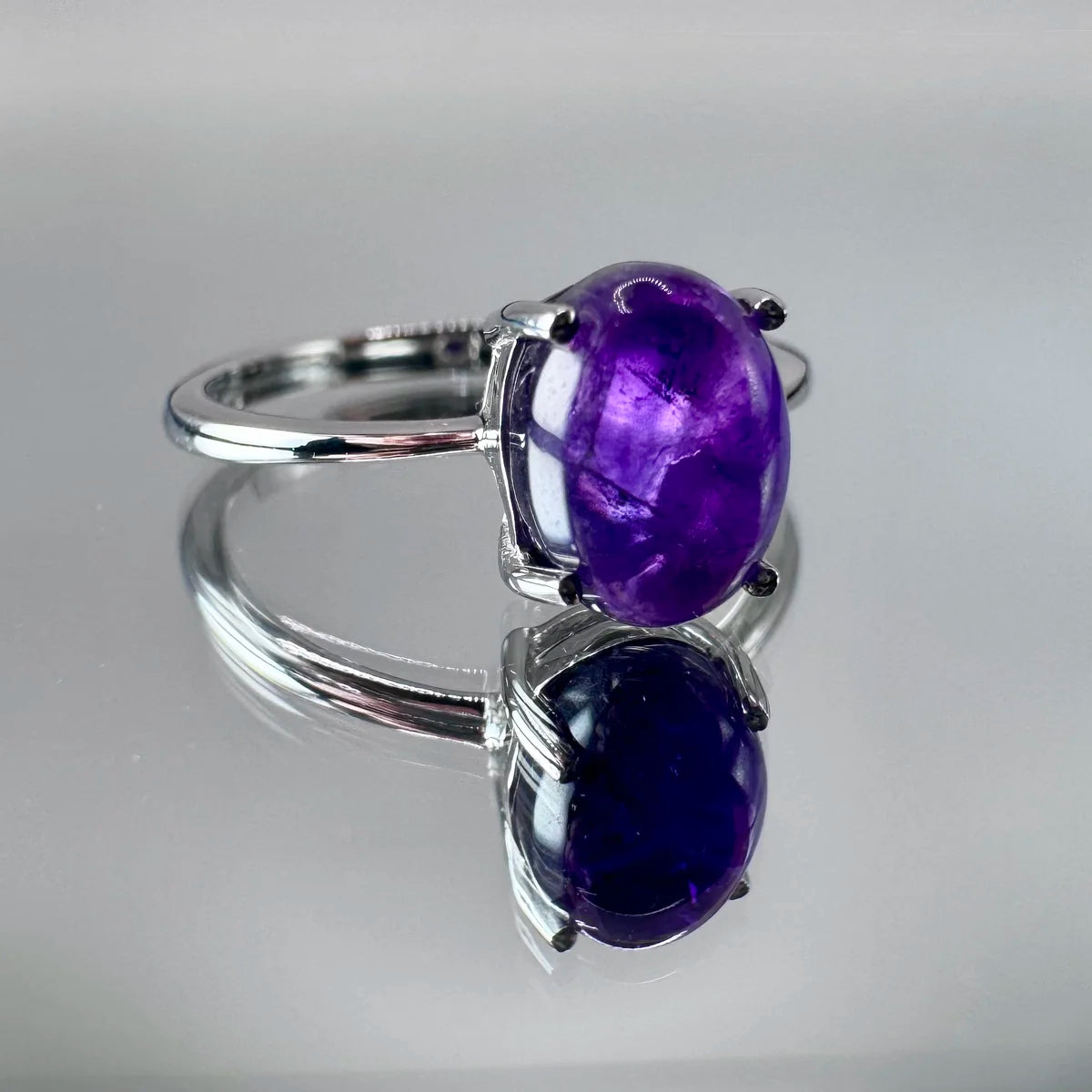 Gros plan sur la bague pierre d'améthyste taille cabochon, posée sur un miroir, reflets violets profonds.